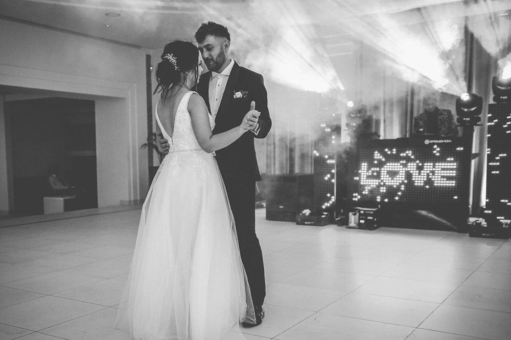 Para młoda w pierwszym tańcu, oświetlenie sceniczne z napisem LOVE w tle, DJ za konsoletą.