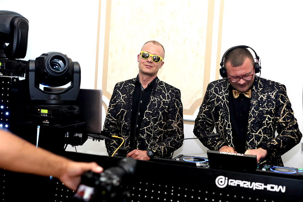 Dwóch DJ-ów w czarno-złotych marynarkach za konsoletą, oświetlenie sceniczne w tle, ujęcie zza ramienia fotografa.