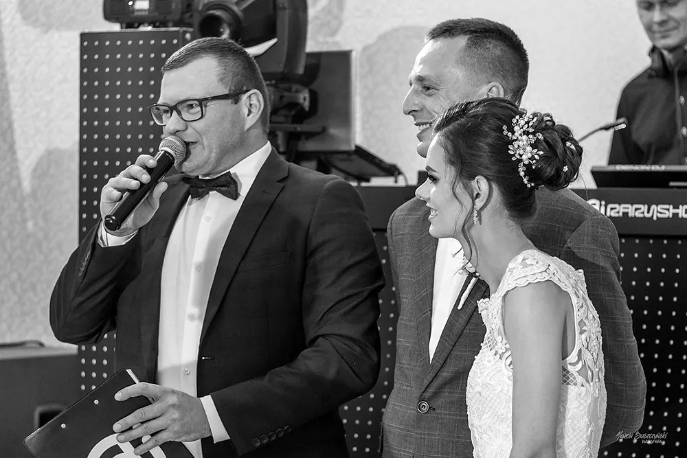 Czarno-białe zdjęcie wodzireja w smokingu z mikrofonem, obok uśmiechnięta para młoda podczas uroczystości weselnej, w tle sprzęt DJ-ski.
