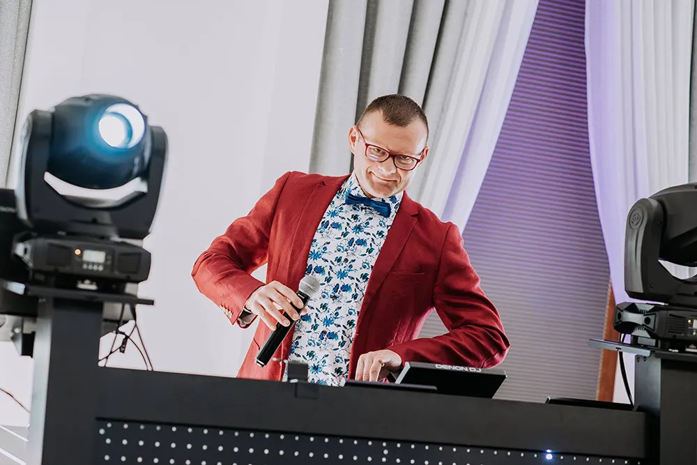 Uśmiechnięty konferansjer w bordowej marynarce, kwiecistej koszuli i muszce, trzymający mikrofon przy stanowisku DJ-a z oświetleniem scenicznym.