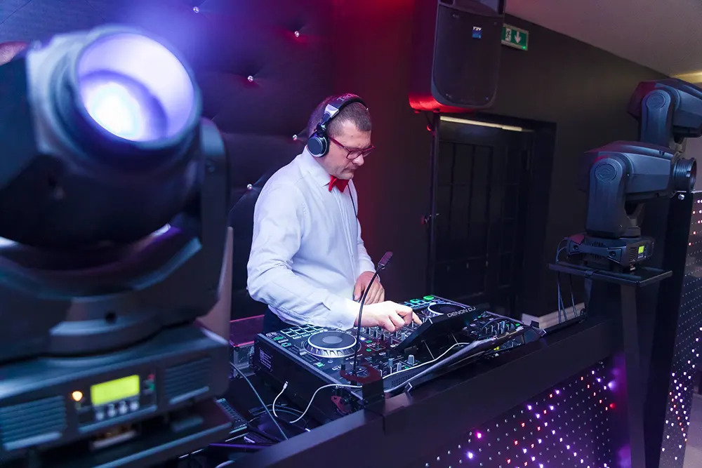 DJ w czerwonej muszce miksuje muzykę na konsolecie Denon DJ, oświetlony reflektorami i światłami LED.