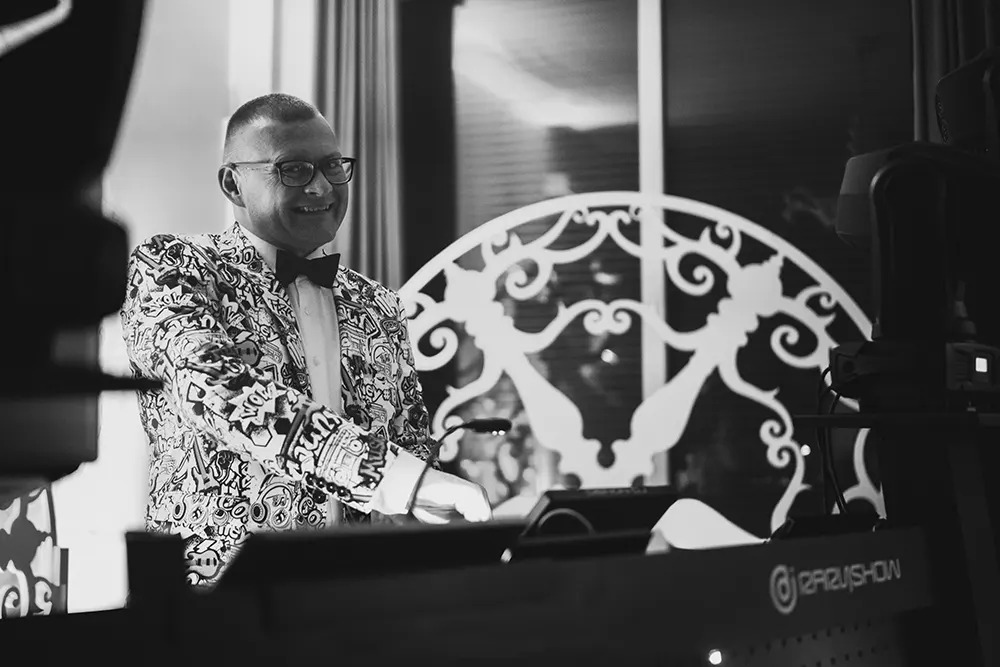 Uśmiechnięty DJ w czarno-białym wzorzystym garniturze i muszce, obsługujący sprzęt nagłośnieniowy na tle dekoracyjnej, ażurowej ścianki.