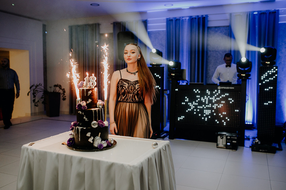 Uśmiechnięta dziewczyna w sukience obok trzypiętrowego tortu urodzinowego z fontannami iskier, w tle DJ na stanowisku z oświetleniem punktowym.