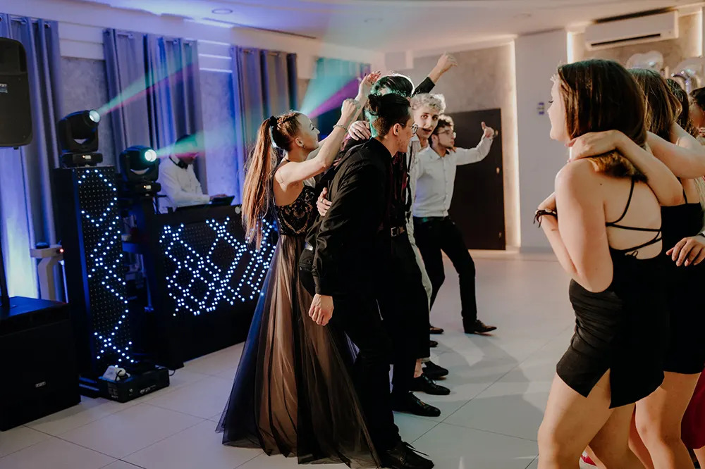 Rozbawieni goście weselni tańczą w rytm muzyki, a DJ za konsolą i oświetlenie tworzą imprezową atmosferę w Kaliszu.