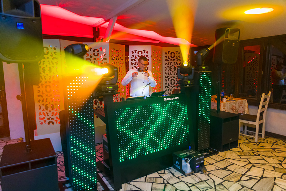 DJ w białej koszuli i okularach, za konsolą z zielonymi diodami, otoczony nagłośnieniem i oświetleniem scenicznym podczas imprezy.