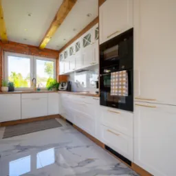 Jasna kuchnia z białymi szafkami, drewnianymi blatami i ceglaną ścianą, widok na zewnątrz przez okno, marmurowe płytki na podłodze.