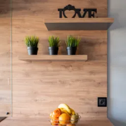 Fragment kuchni z blatem drewnopodobnym, na którym stoi koszyk z owocami, a na półkach powyżej widoczne trzy doniczki z trawą i dekoracyjny napis LOVE, całość na tle drewnianej ściany.