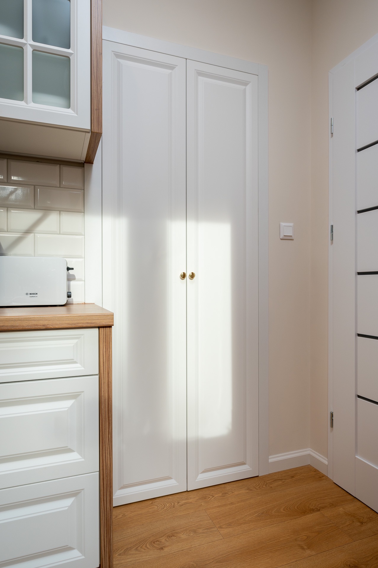 Białe, dwuskrzydłowe drzwi wnękowe z okrągłymi, złotymi gałkami, w tle fragment zabudowy kuchennej z białymi frontami i drewnianym blatem, widoczny również fragment białych drzwi z czarnymi...