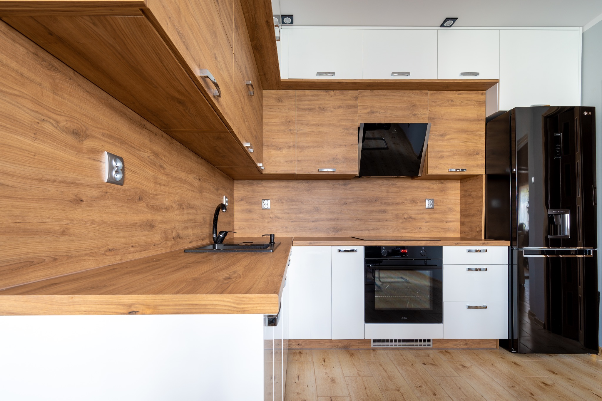 Nowoczesna kuchnia z drewnianymi frontami szafek górnych i blatem, białymi szafkami dolnymi oraz czarnym sprzętem AGD: piekarnikiem i lodówką.