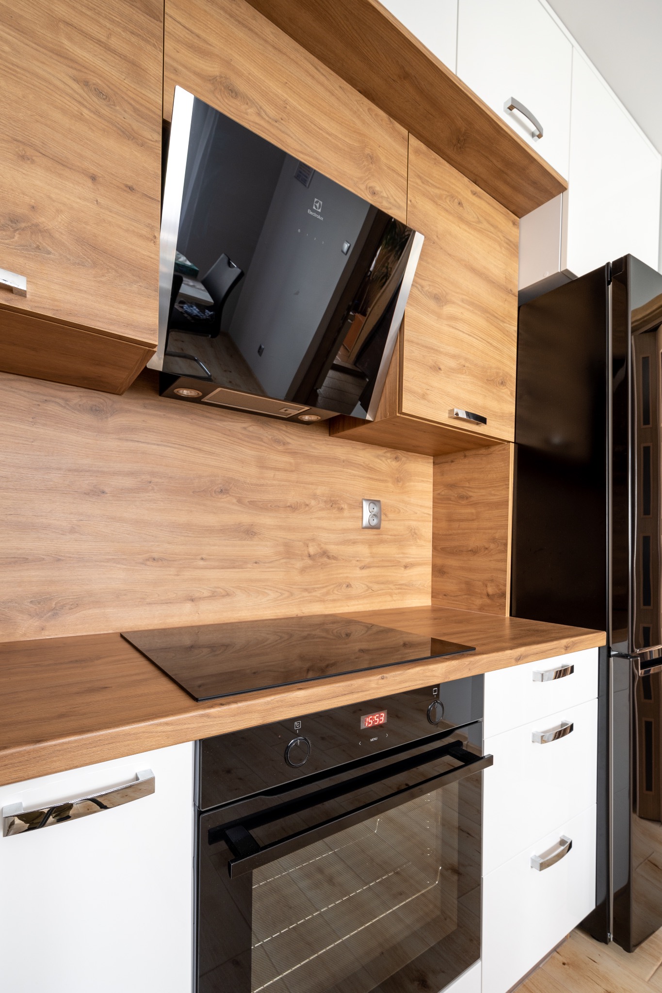 Nowoczesna kuchnia z drewnianymi szafkami górnymi i blatem, białymi szafkami dolnymi, czarną płytą indukcyjną i piekarnikiem; widoczny fragment lodówki.