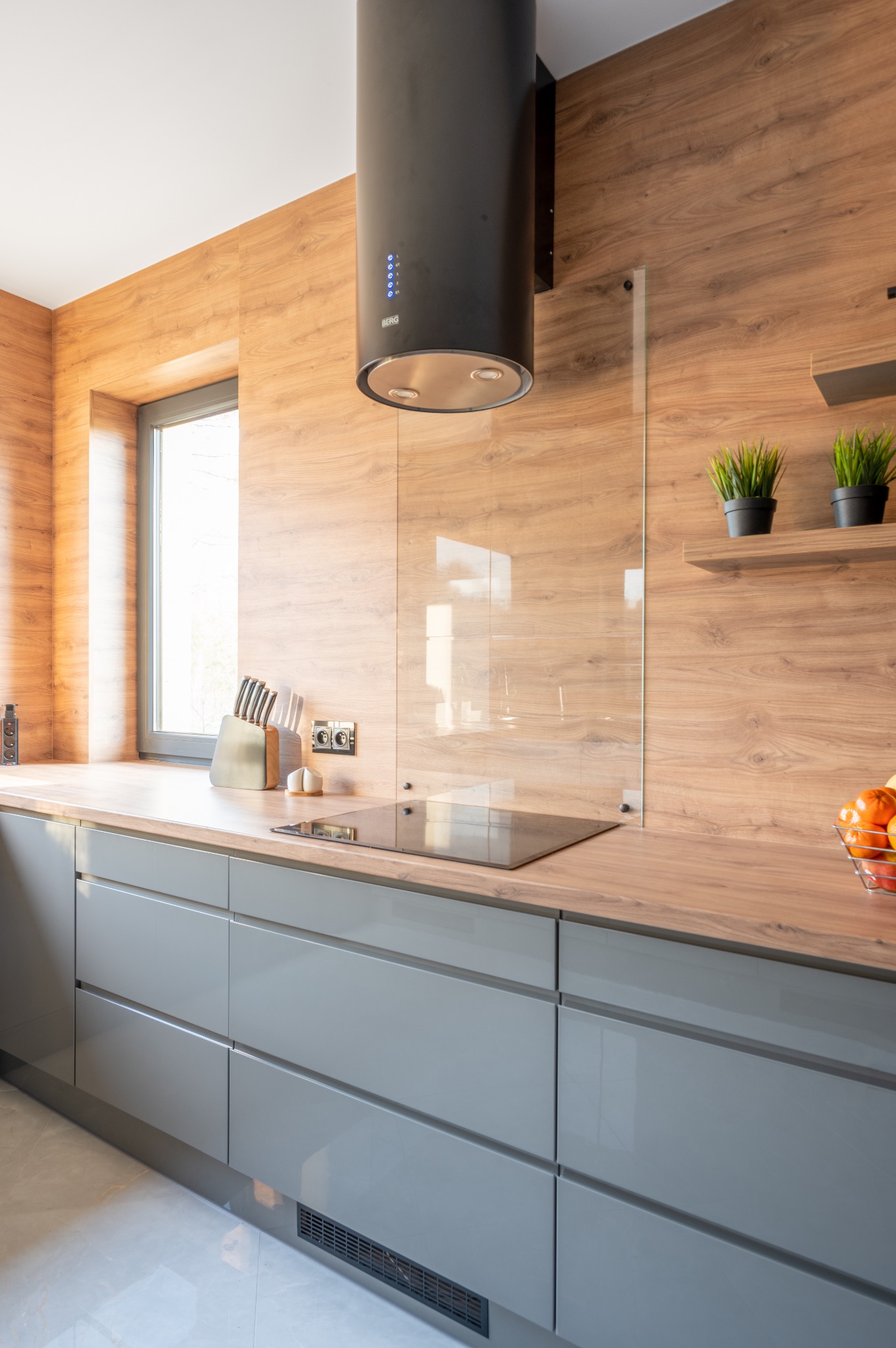 Nowoczesna kuchnia z szarymi, gładkimi frontami szafek, drewnianym blatem i okładziną ścienną, cylindrycznym okapem nad płytą indukcyjną oraz szklanym panelem ochronnym.