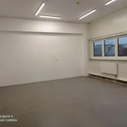 Puste, jasne pomieszczenie biurowe z trzema oknami PCV, grzejnikiem i szarą posadzką, przygotowane do malowania.