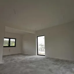 Surowe wnętrze po malowaniu, z widocznymi wystającymi przewodami elektrycznymi, szarym betonowym podłożem i nowymi oknami z czarnymi ramami.