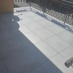 Wyłożony jasnymi płytkami balkon w Gniewie z ozdobną, metalową balustradą i cieniem rzucanym przez budynek, widoczny mop do czyszczenia podłogi.