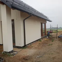 Elewacja domu jednorodzinnego w trakcie malowania, widoczne rusztowanie, pracownik z telefonem oraz fragment pola w tle.