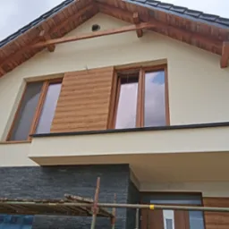 Dwukondygnacyjny dom z ocieploną elewacją w kolorze kremowym, z drewnianymi elementami dekoracyjnymi na szczycie i panelami imitującymi drewno przy oknach, oraz wykończeniem z szarego kamienia...