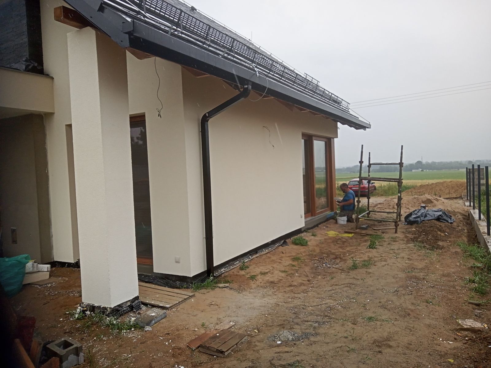 Elewacja domu jednorodzinnego w trakcie malowania, widoczne rusztowanie, pracownik z telefonem oraz fragment pola w tle.