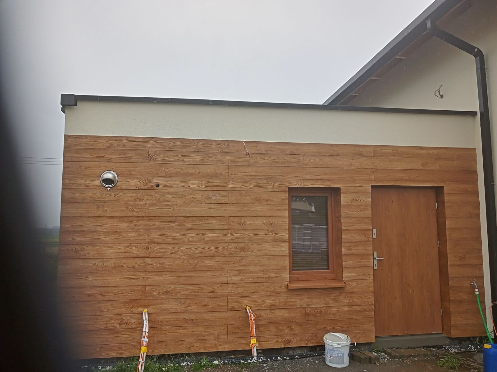 Nowoczesny budynek gospodarczy z elewacją imitującą drewno, widoczne okno, drzwi i wentylacja, na dole leżą zwinięte kable i wiadro z farbą.