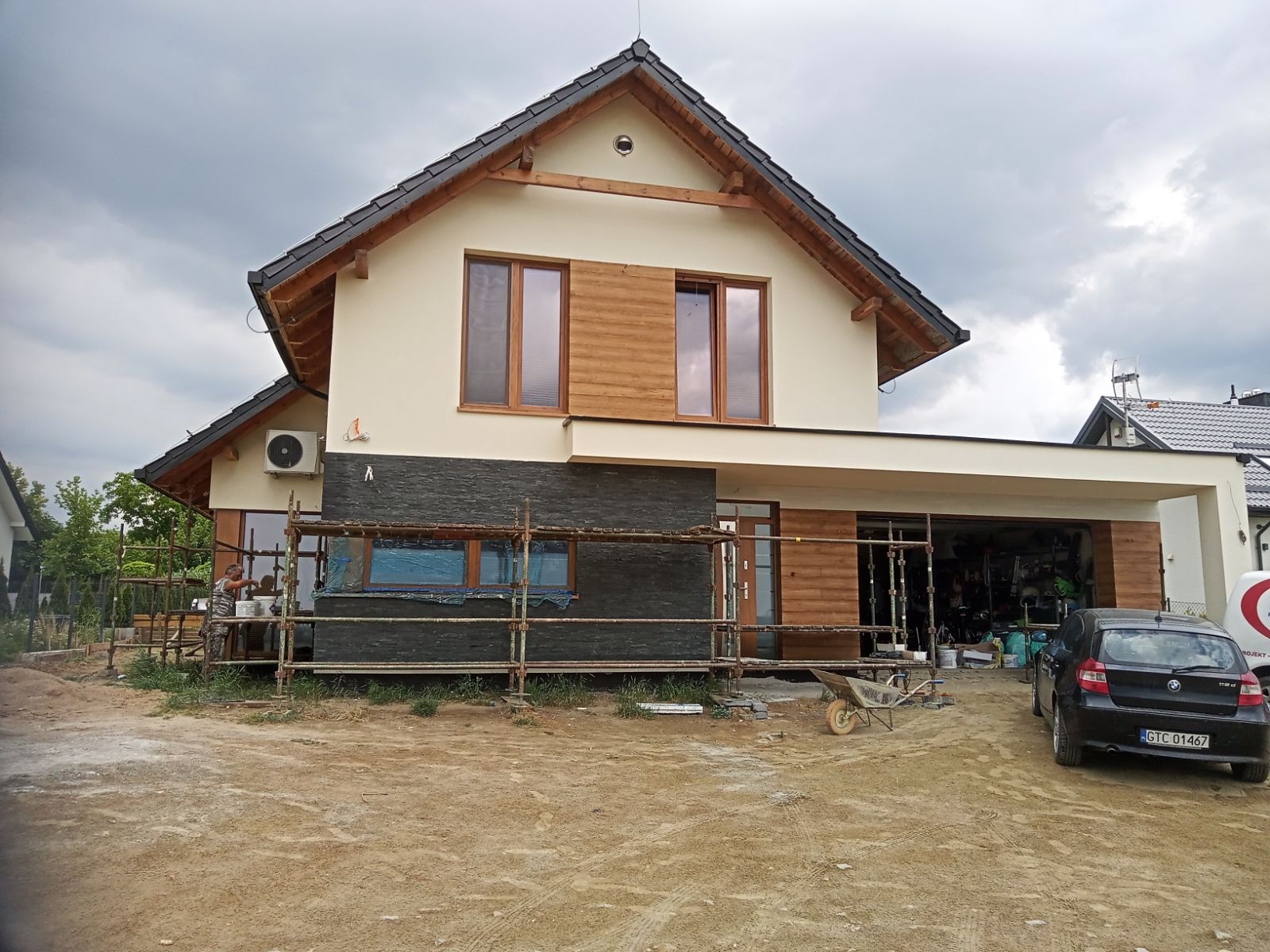 Dwukondygnacyjny dom z jasną elewacją i elementami drewnianymi, w trakcie prac wykończeniowych elewacji, widoczne rusztowania, pracownik przy elewacji oraz zaparkowany samochód osobowy przed...