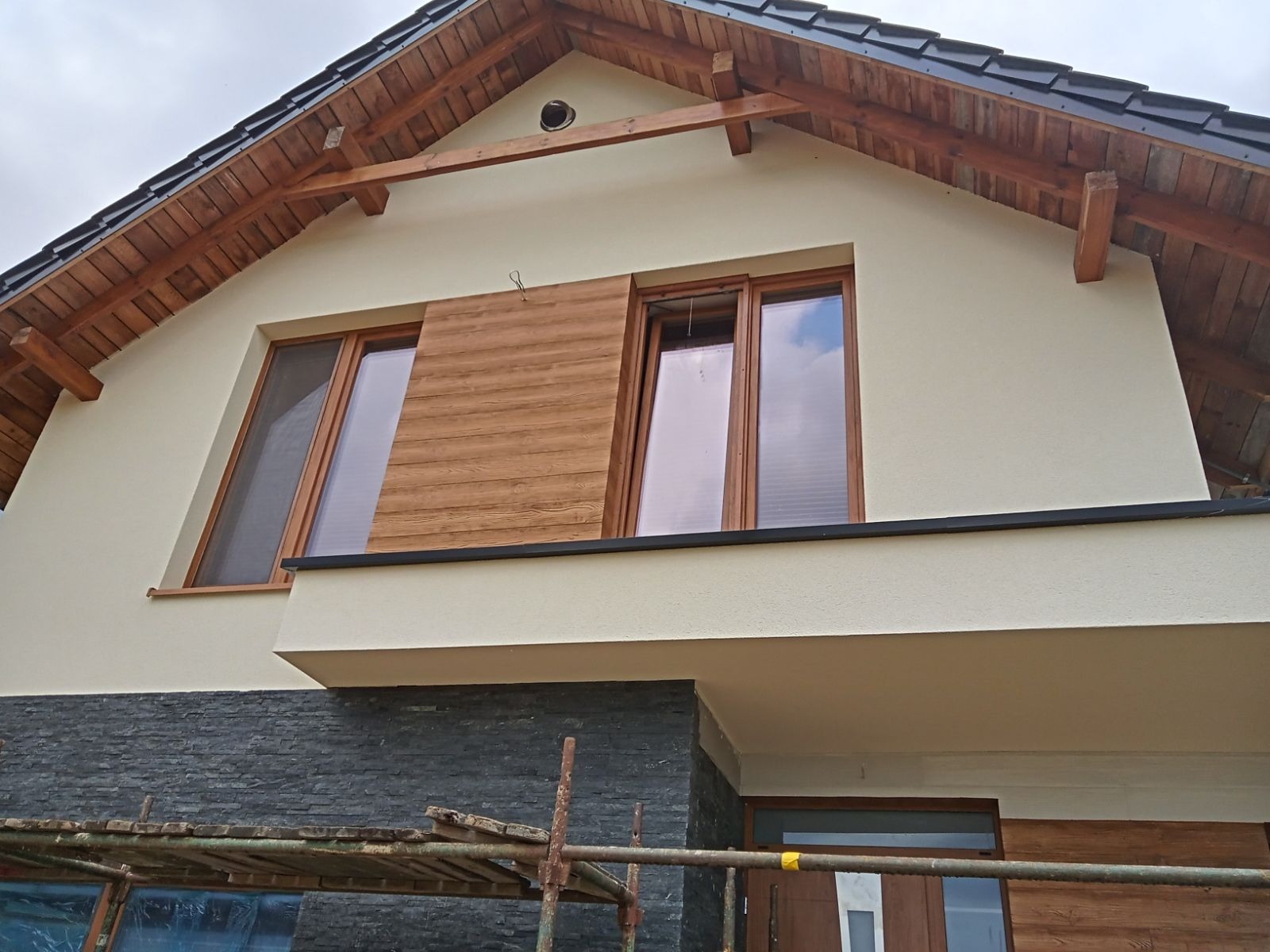 Dwukondygnacyjny dom z ocieploną elewacją w kolorze kremowym, z drewnianymi elementami dekoracyjnymi na szczycie i panelami imitującymi drewno przy oknach, oraz wykończeniem z szarego kamienia...