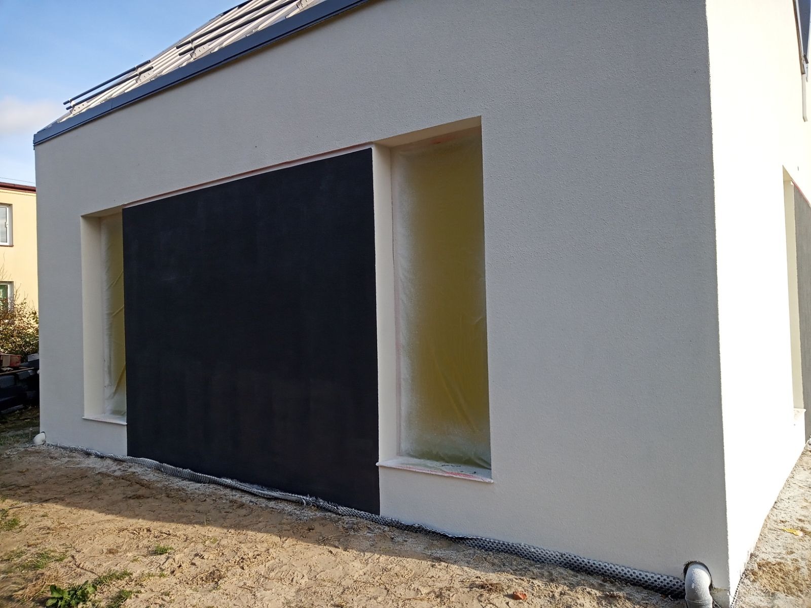 Fragment elewacji budynku w trakcie prac: świeżo otynkowana, jasna ściana z częściowo zamaskowanym oknem i czarną, przygotowaną powierzchnią pod dalsze prace wykończeniowe.