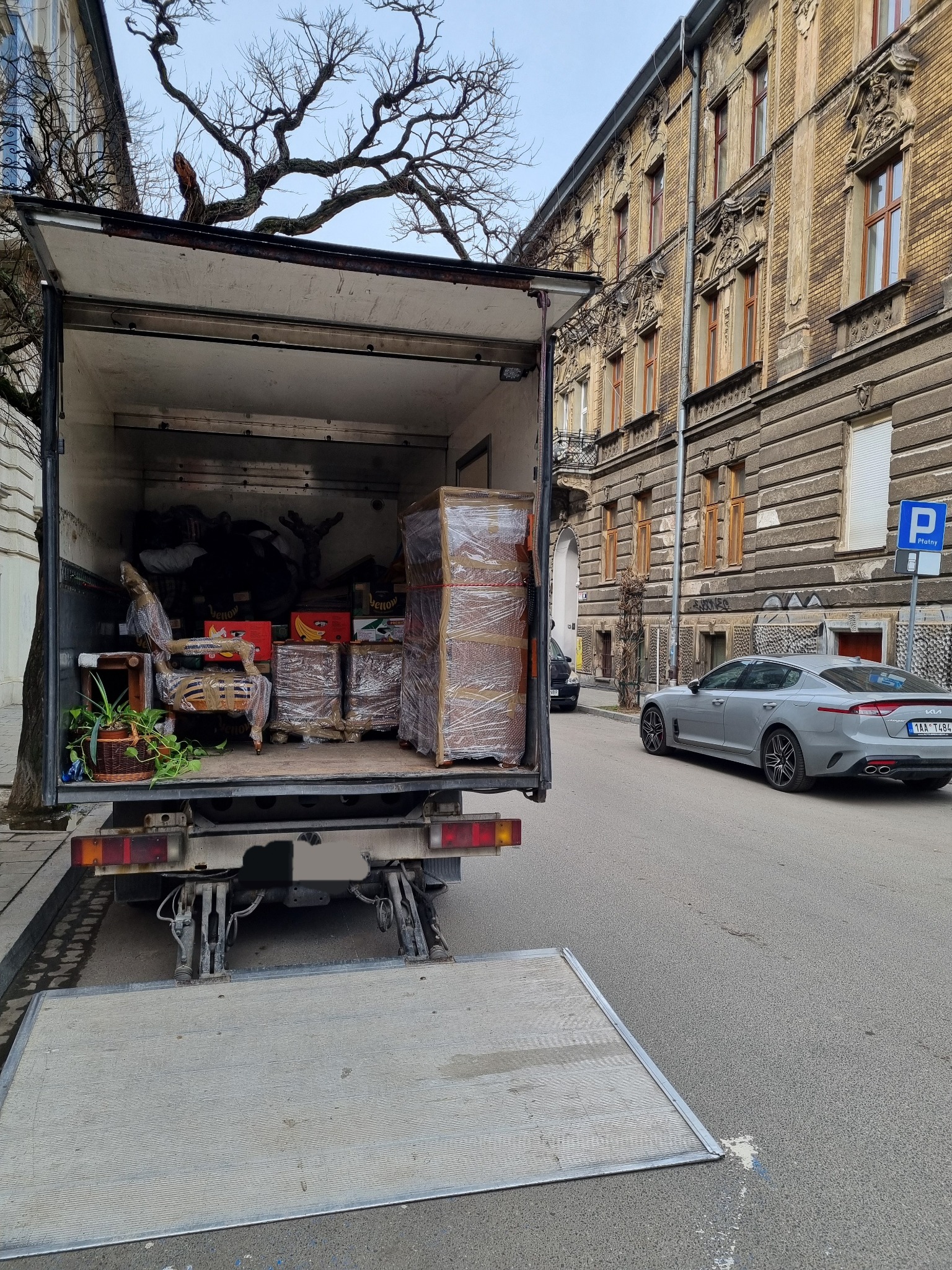 Otwarta paka ciężarówki wypełniona meblami i kartonami zabezpieczonymi folią stretch, przygotowanymi do transportu na tle kamienicy i zaparkowanego szarego samochodu osobowego.