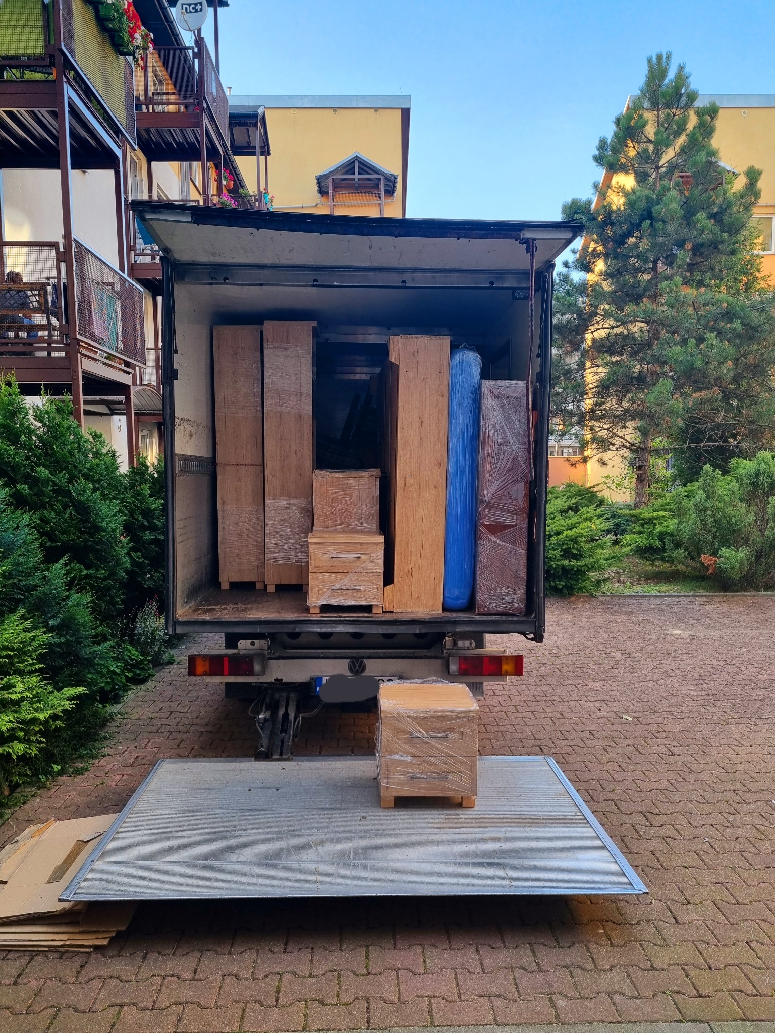 Widok z tyłu otwartego samochodu dostawczego z windą załadowczą, wypełnionego meblami zabezpieczonymi folią stretch, na tle budynku mieszkalnego i zieleni.