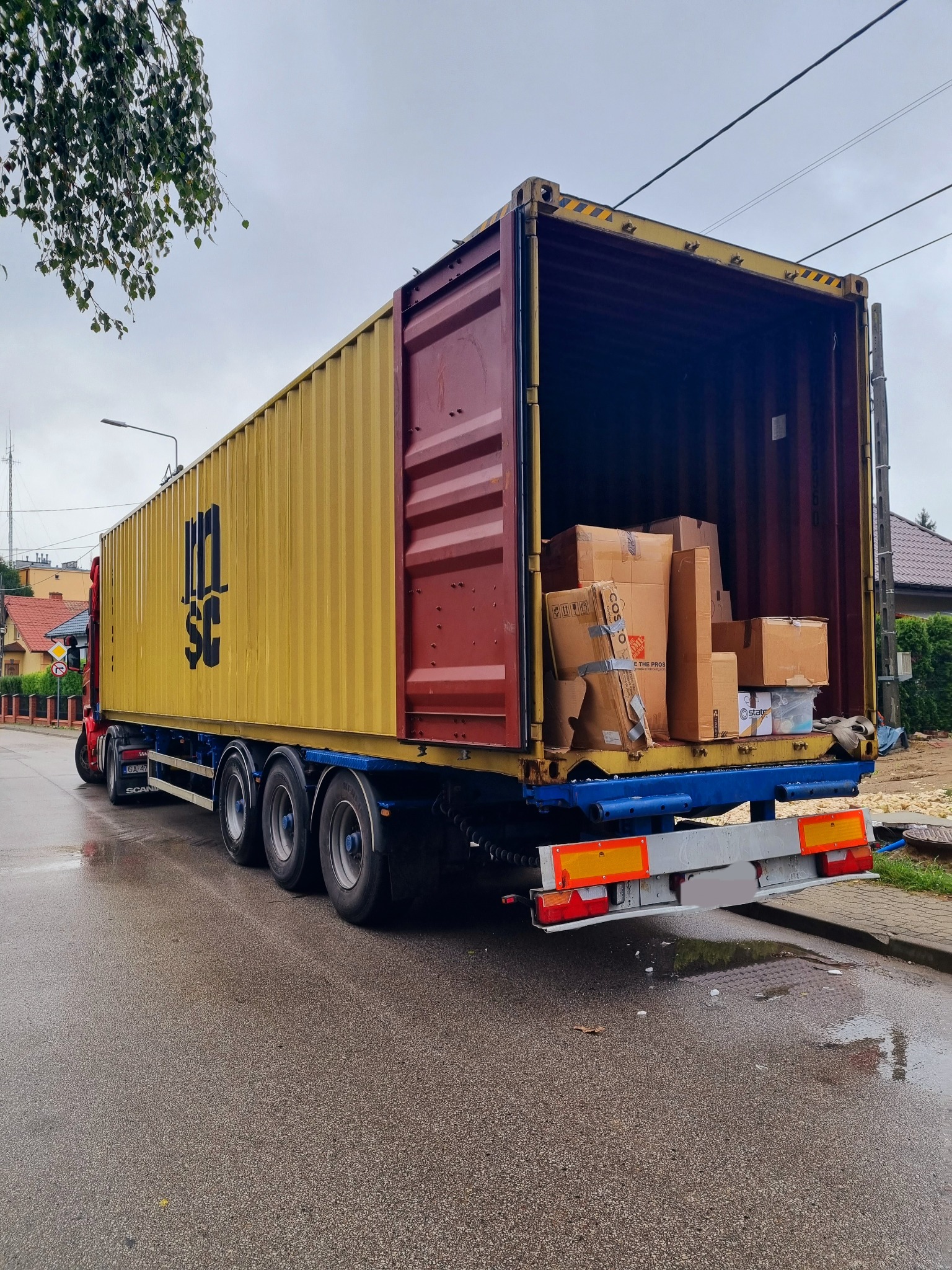 Otwarty kontener na naczepie ciężarówki, wypełniony kartonami z różnymi przedmiotami, na tle ulicy w pochmurny dzień.