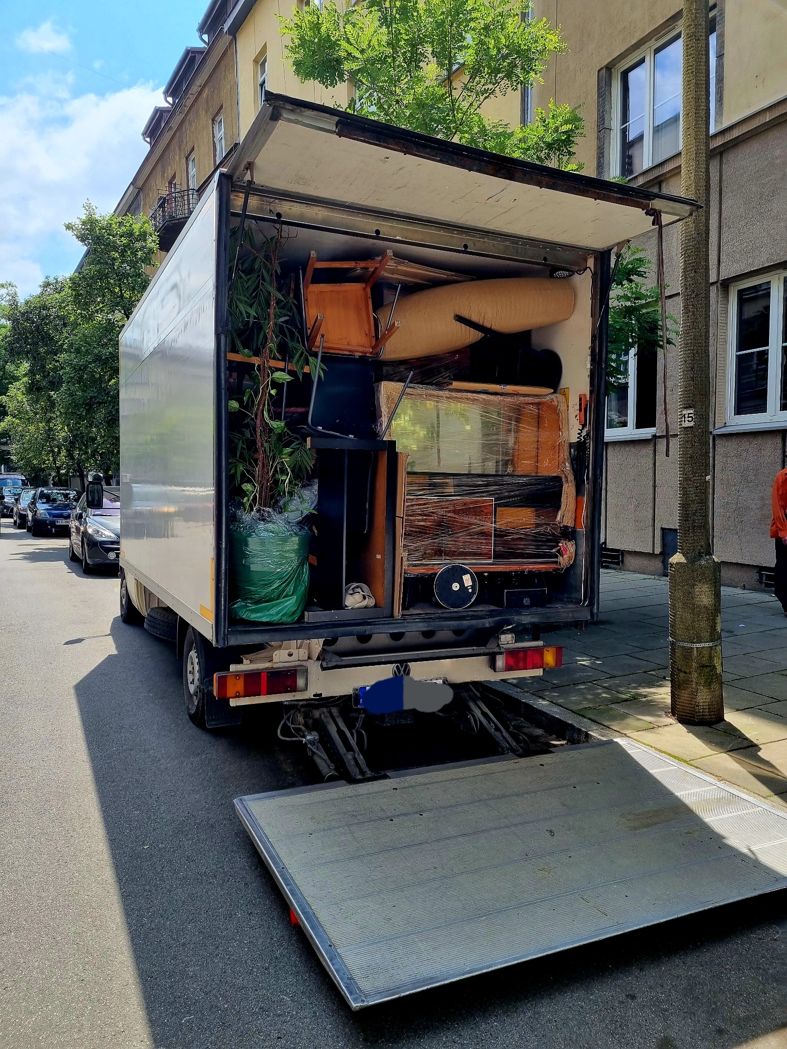 Otwarta paka samochodu dostawczego załadowana meblami, rośliną doniczkową w zielonym worku i innymi przedmiotami, gotowa do transportu podczas przeprowadzki w mieście.