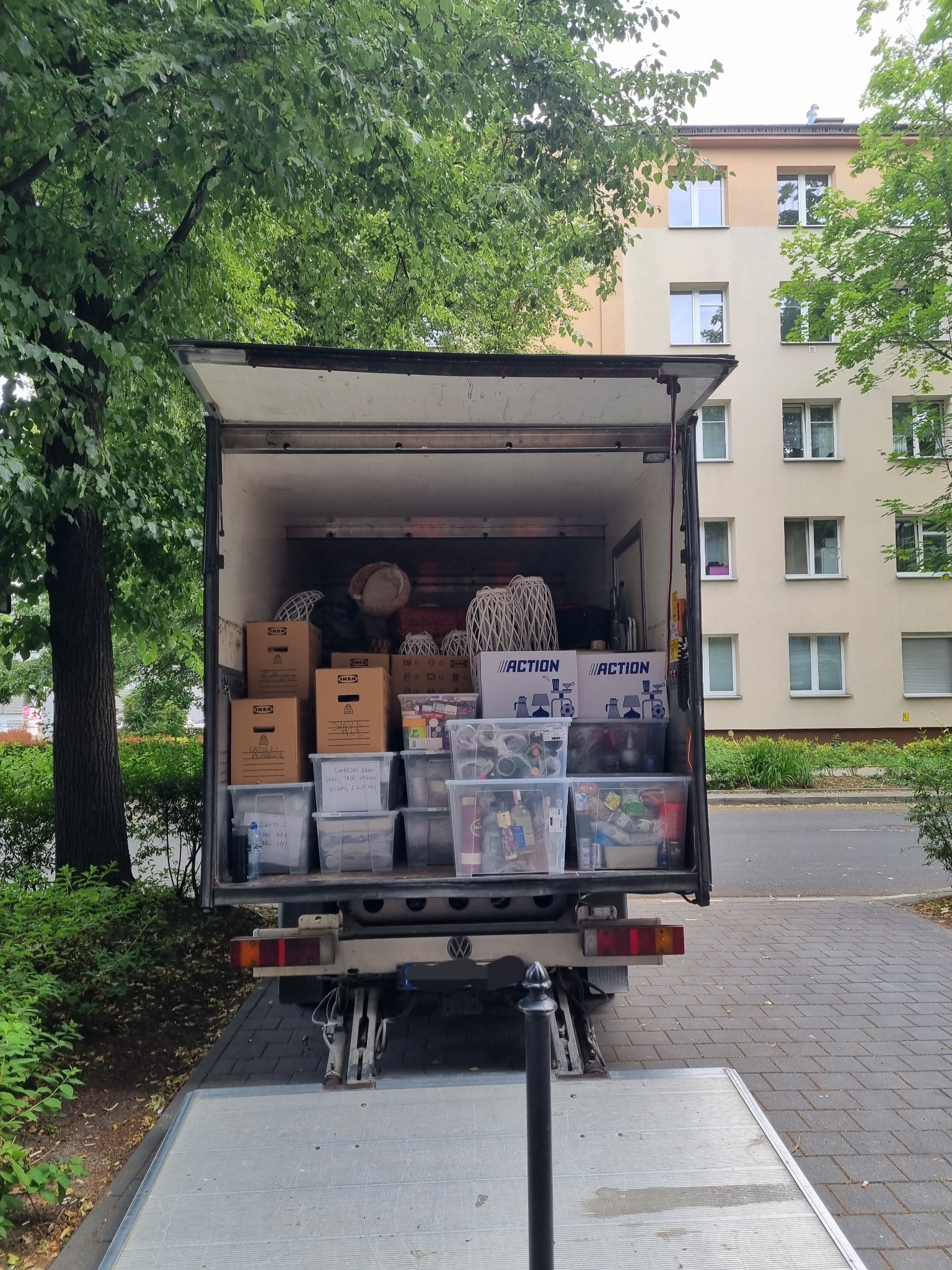 Otwarta przestrzeń ładunkowa samochodu dostawczego wypełniona kartonami, plastikowymi pojemnikami i innymi przedmiotami podczas przeprowadzki, z widocznymi napisami na kartonach i pojemnikach...