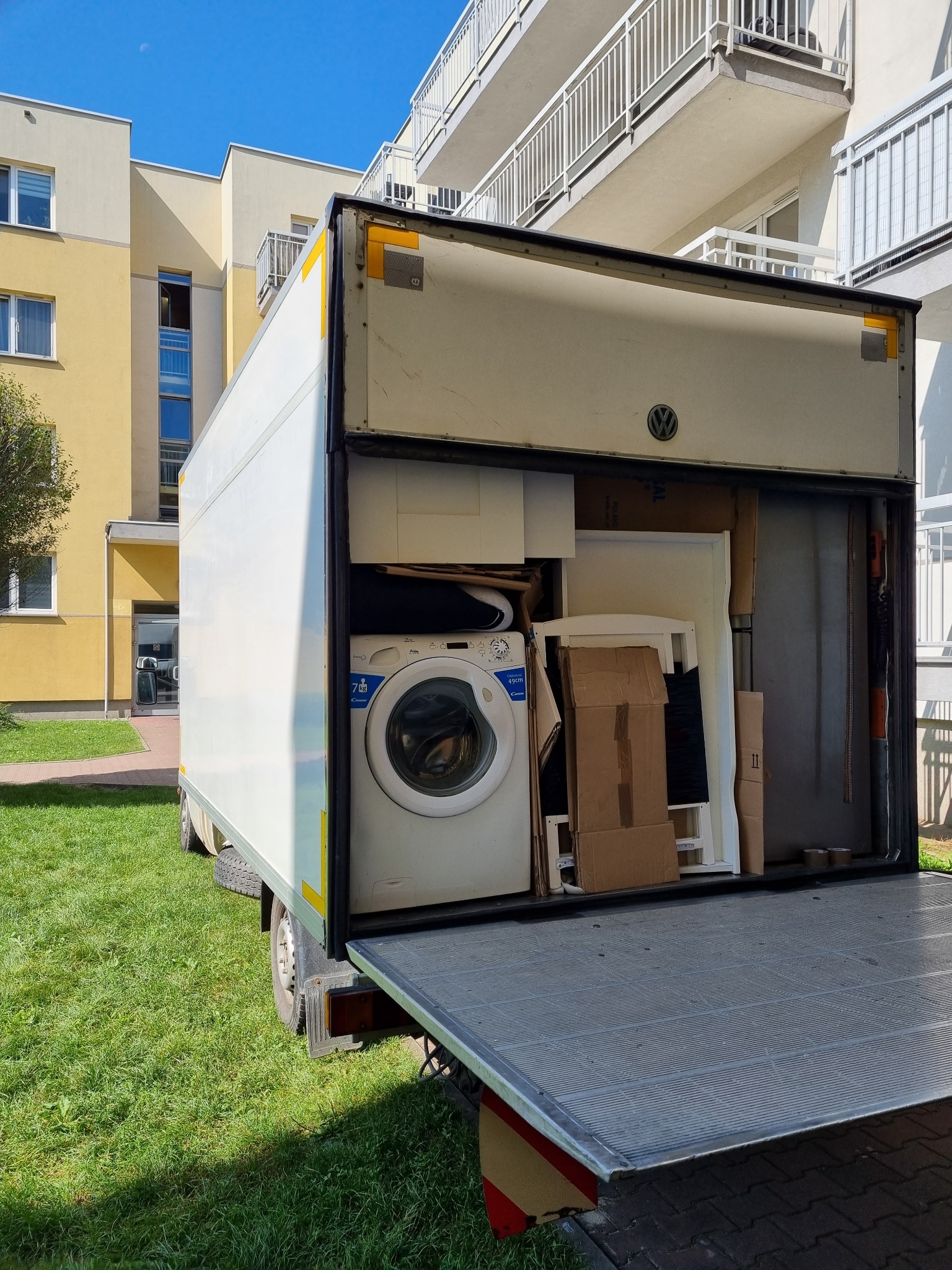 Otwarta paka samochodu dostawczego wypełniona sprzętem AGD, meblami i kartonami podczas przeprowadzki, na tle bloku mieszkalnego z balkonami.