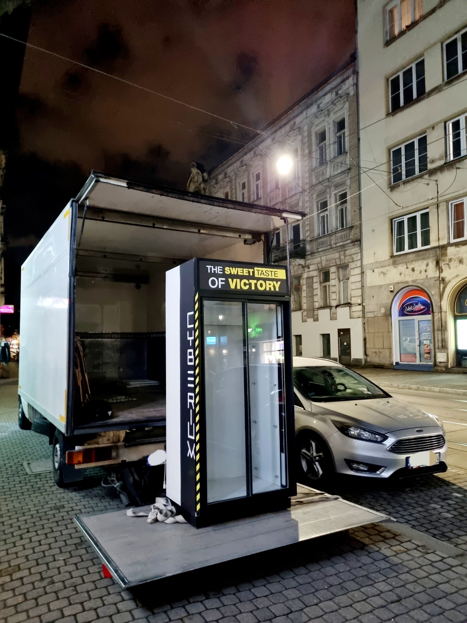 Nocny załadunek chłodziarki z napisem 'The Sweet Taste of Victory' do otwartej furgonetki na tle miejskiej ulicy z zaparkowanym srebrnym Fordem Focusem.