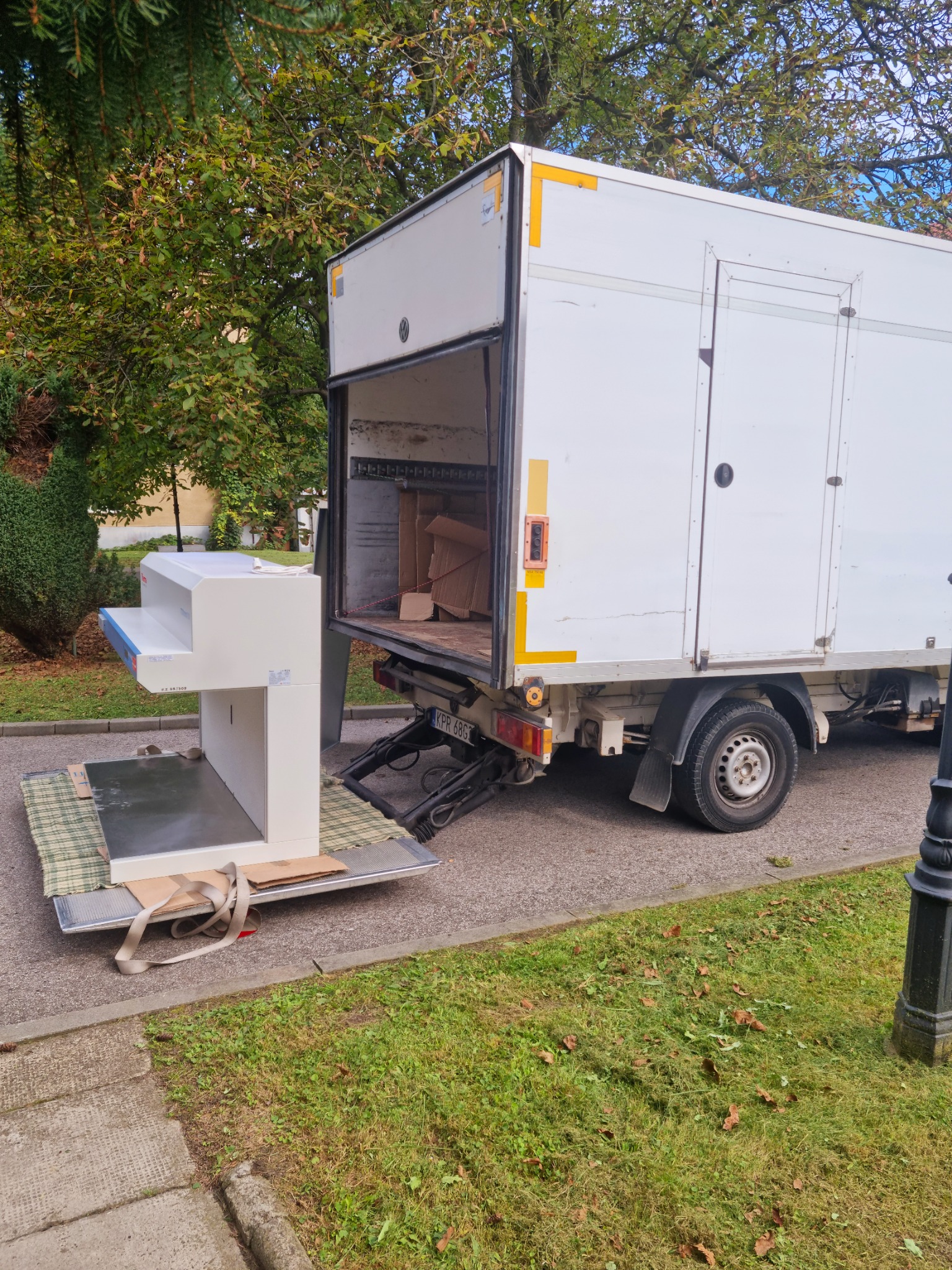Białe pianino zabezpieczone pasami na platformie windy załadunkowej, gotowe do wjechania do białej furgonetki transportowej na tle zieleni.