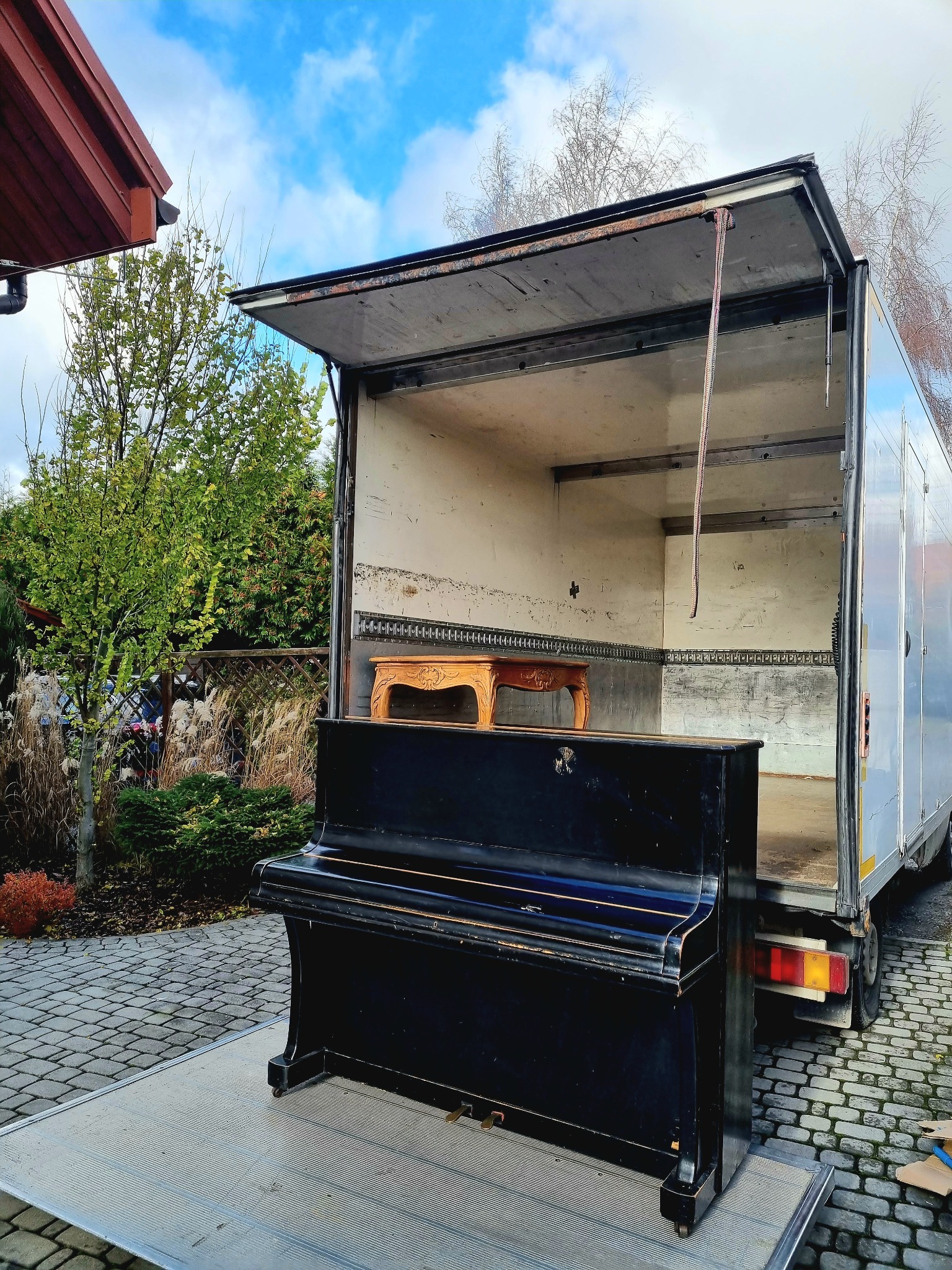 Czarne pianino na platformie załadunkowej otwartego samochodu dostawczego, w tle widoczny ozdobny stolik i zielona roślinność.