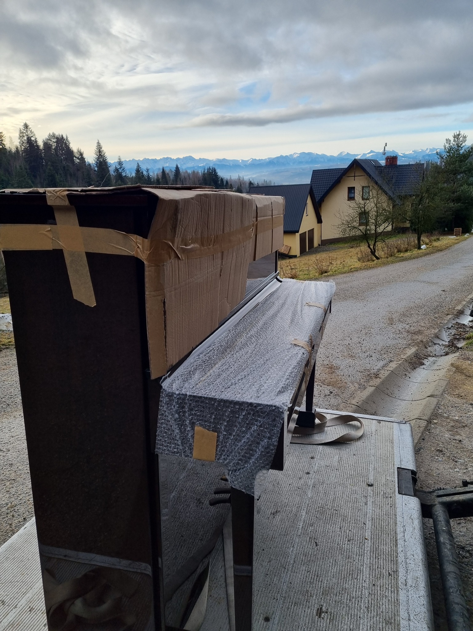 Pianino zabezpieczone kartonem i folią bąbelkową, transportowane na platformie samochodu dostawczego; w tle widok na domy i pasmo górskie.