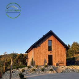 Beskid Haus - Nowoczesny dom z drewna o prostej bryle, z dużym oknem nad wejściem, otoczony kamieniami i zielenią, w ciepłym świetle zachodzącego słońca.