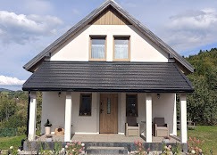 Niewielki, parterowy dom z poddaszem użytkowym w stylu skandynawskim. Biała elewacja, ciemny dach, drewniane elementy dekoracyjne, widok z przodu, zielone otoczenie.