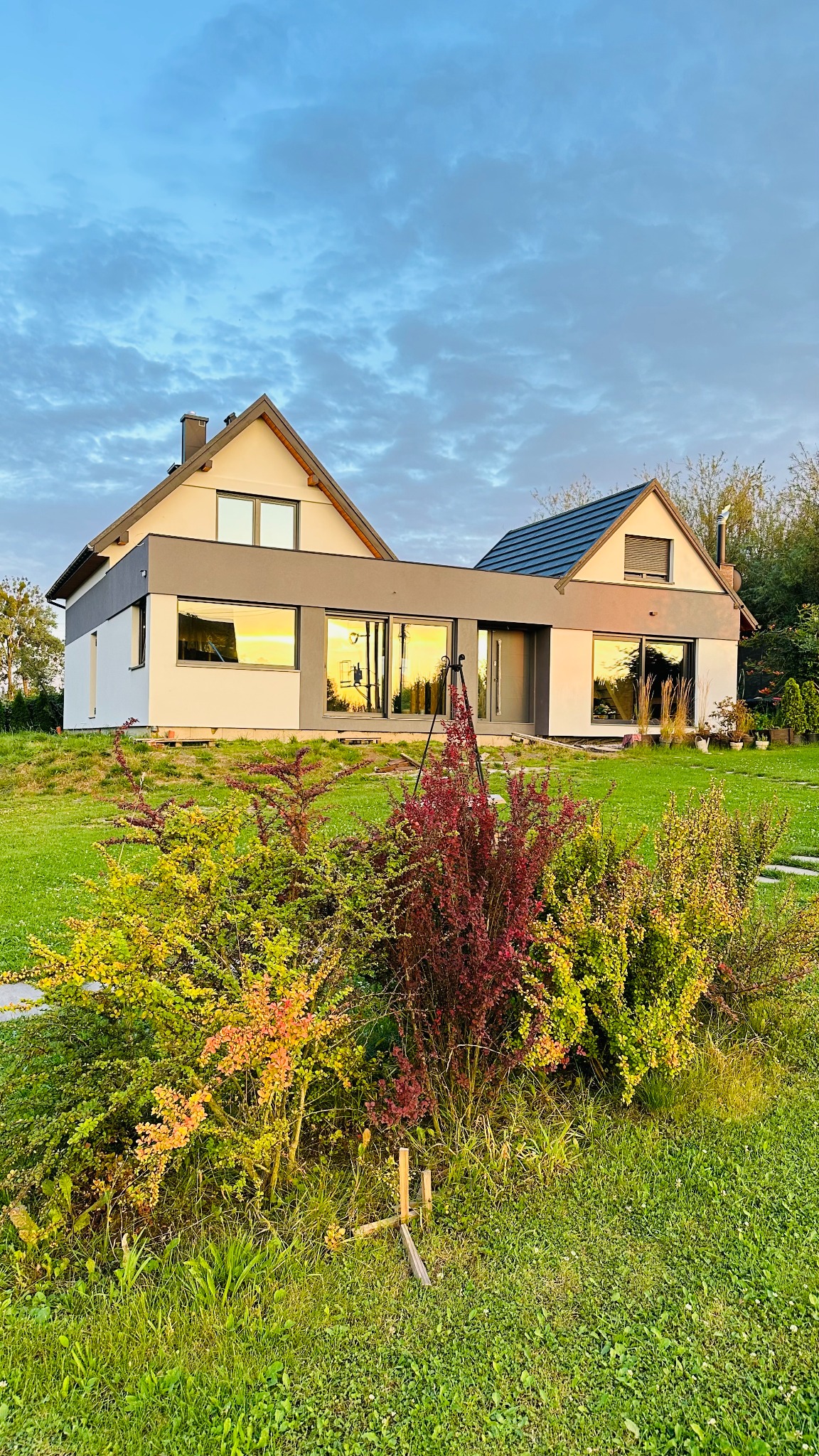 Nowoczesny dom z jasną elewacją i szarymi akcentami, otoczony zielenią. W oknach odbija się zachodzące słońce, a na pierwszym planie kolorowe krzewy.