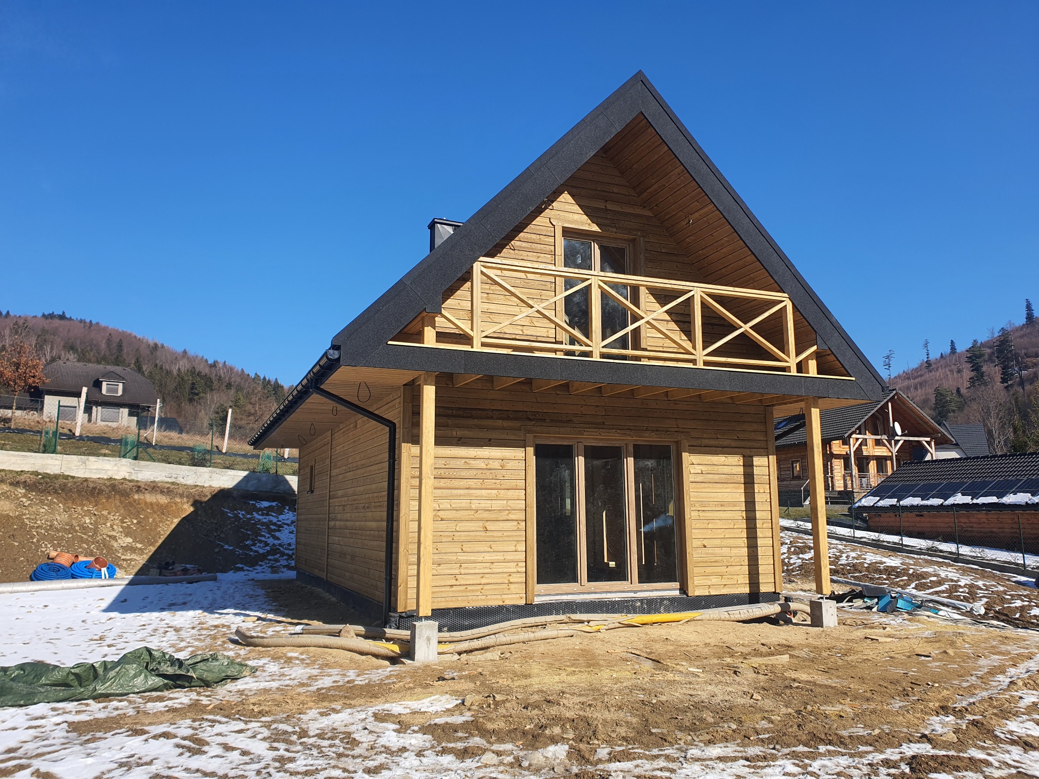 Drewniany dom szkieletowy z balkonem, czarnym dachem i widocznymi elementami konstrukcyjnymi na tle błękitnego nieba i górskiego krajobrazu. Wokół domu rozciąga się teren budowy.