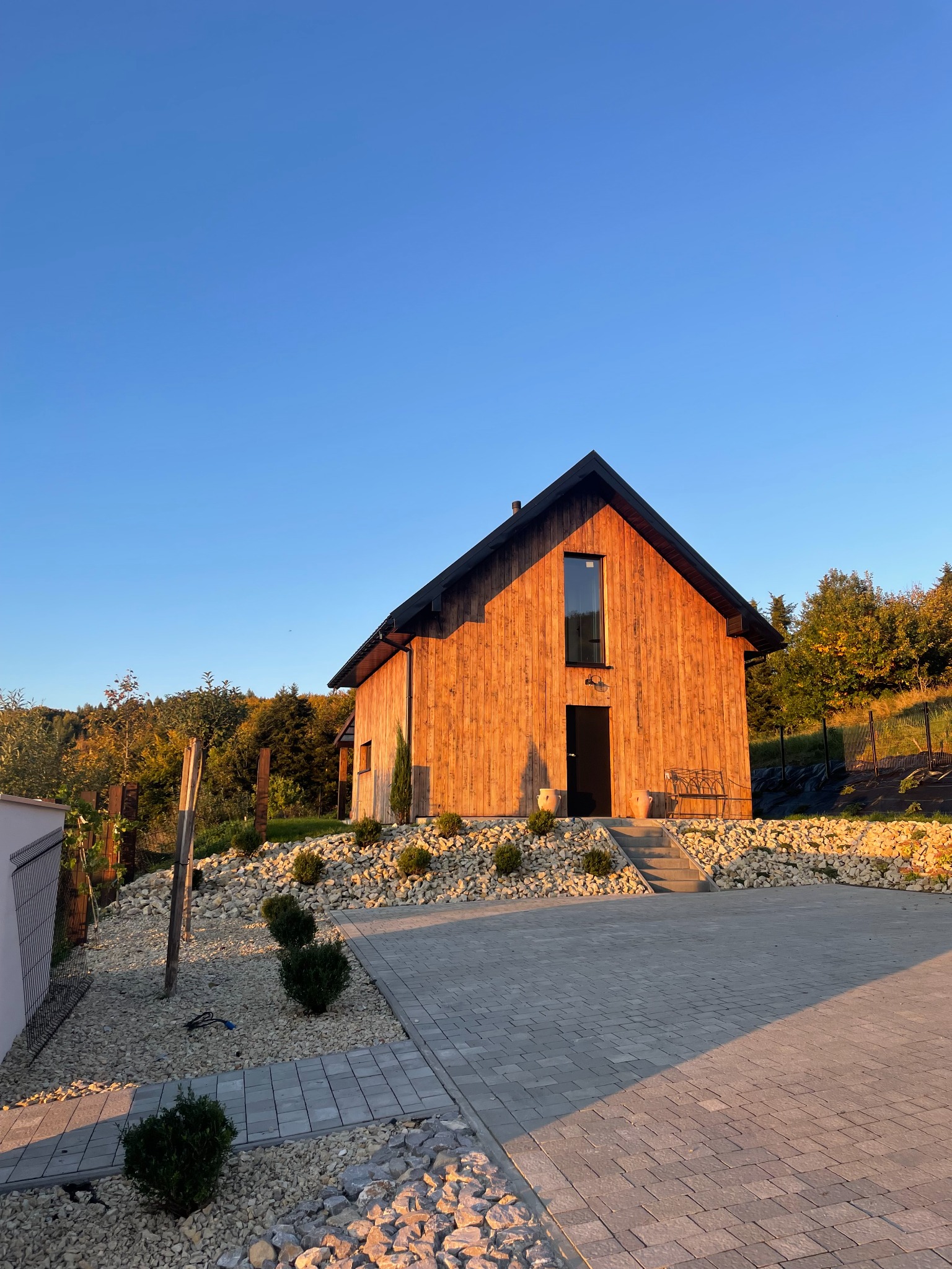 Drewniany dom z czarnym dachem i oknami, otoczony kamieniami i zielenią, oświetlony ciepłym światłem zachodzącego słońca. Widok z dołu.