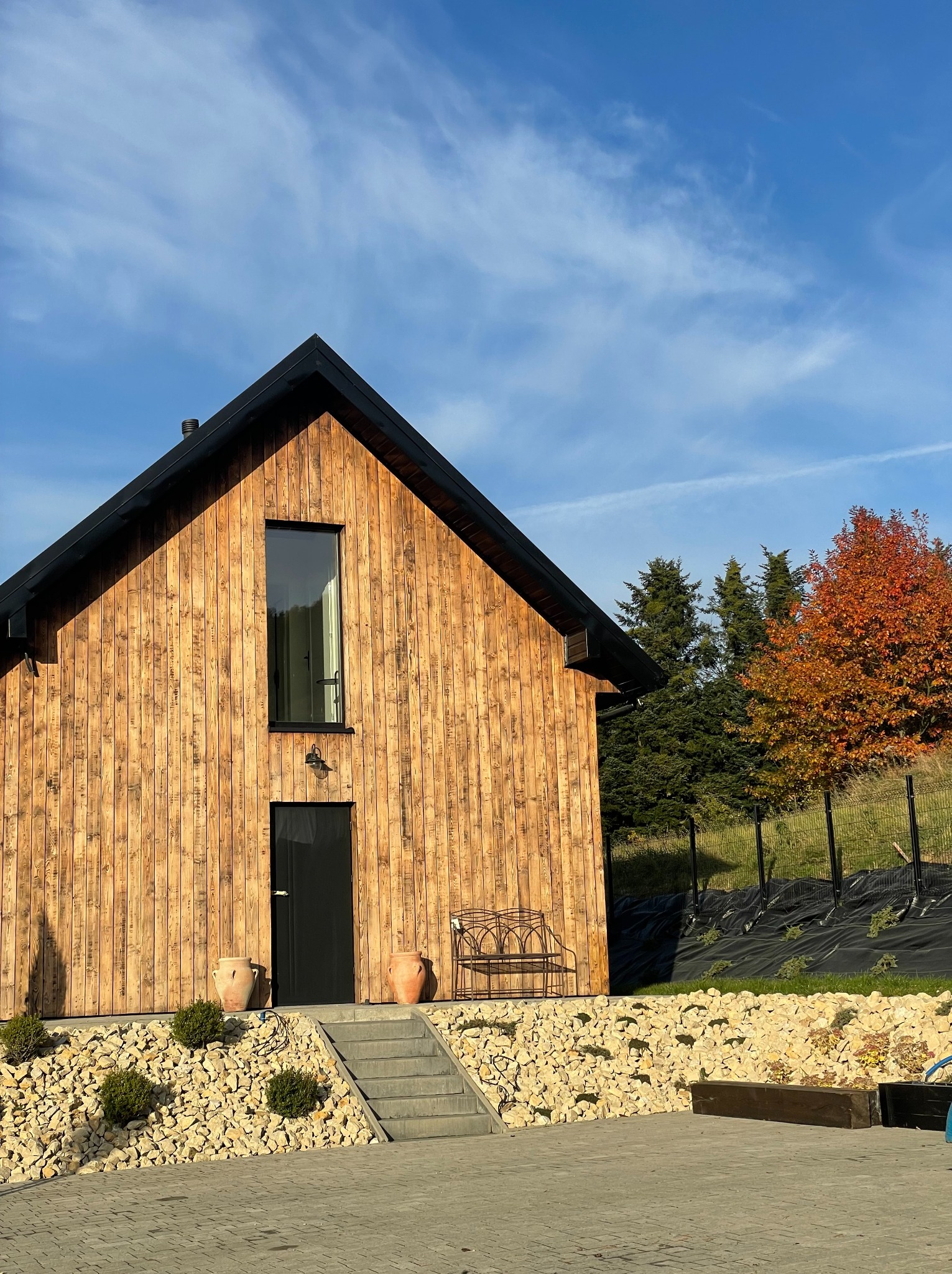 Nowoczesny dom szkieletowy z drewnianą fasadą i czarnym dachem. Uwagę zwraca minimalistyczne okno i otoczenie z kamieni oraz roślin.