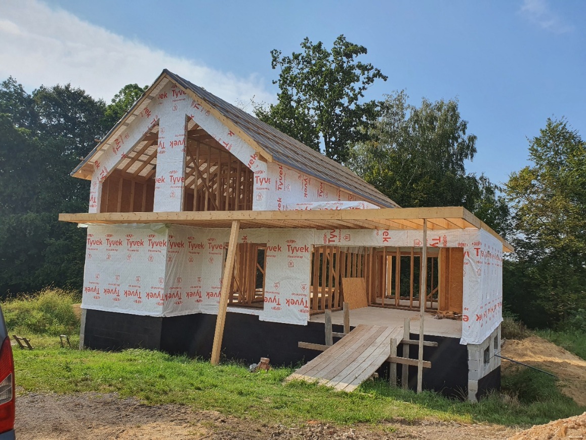 Wznoszenie domu szkieletowego: konstrukcja z drewna, zabezpieczona folią, widok z zewnątrz, w tle zieleń drzew, etap budowy, fundament z bloczków.