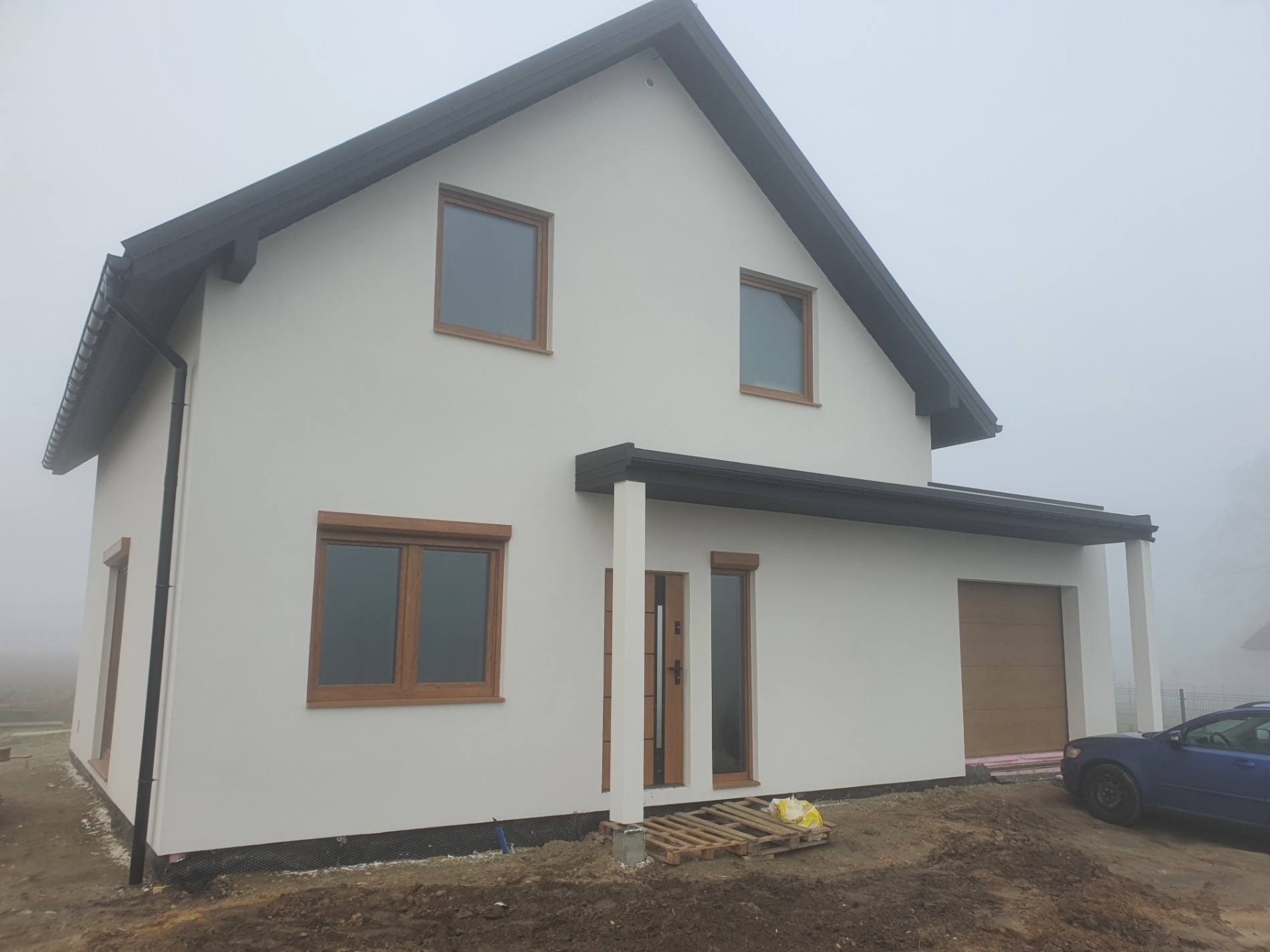 Ukończony dom z białymi ścianami, brązowymi oknami i drzwiami w pochmurny dzień. Widoczna boczna elewacja z garażem i fragment niebieskiego samochodu.