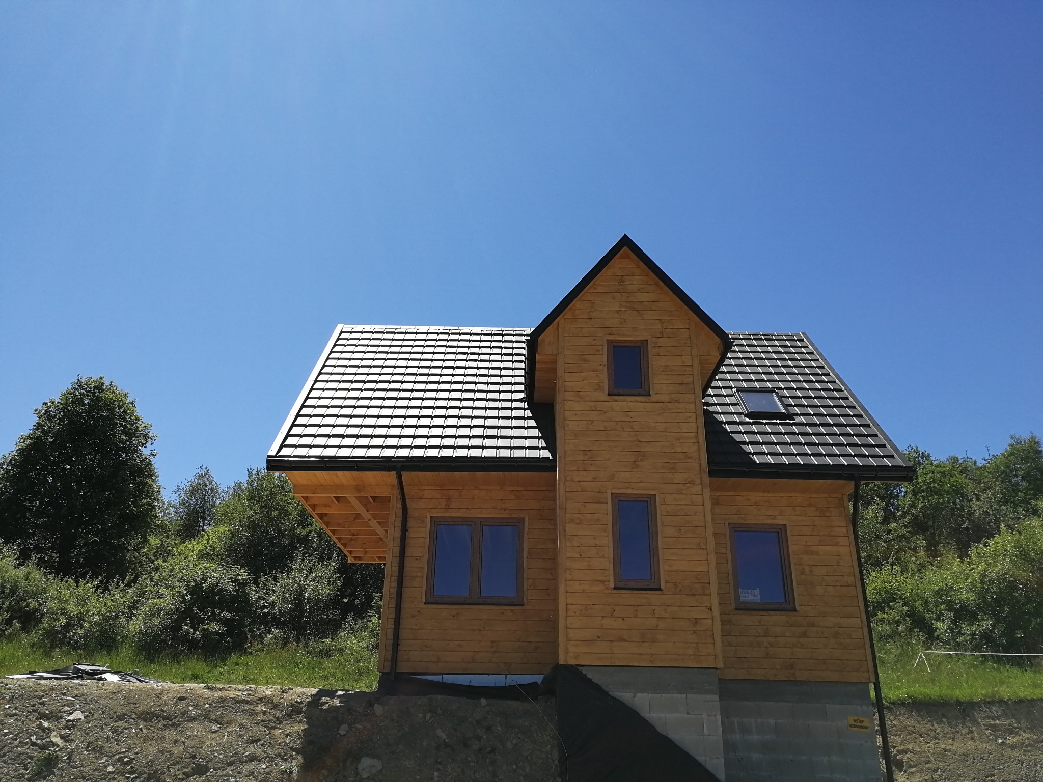 Nowoczesny dom szkieletowy z drewnianą elewacją i ciemną dachówką, widok z dołu, błękitne niebo w tle. Widoczne okna i elementy konstrukcyjne.