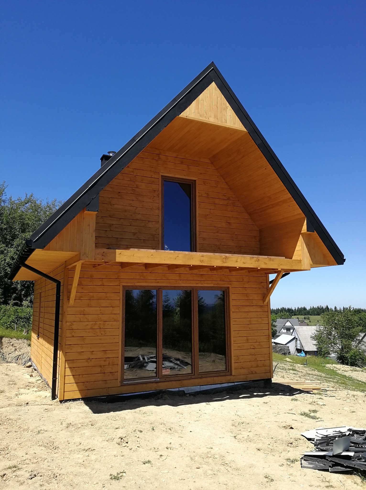 Surowy, drewniany dom szkieletowy z czarnym dachem na tle błękitnego nieba. Widoczne okna i komin. Teren wokół domu przygotowany pod dalsze prace.