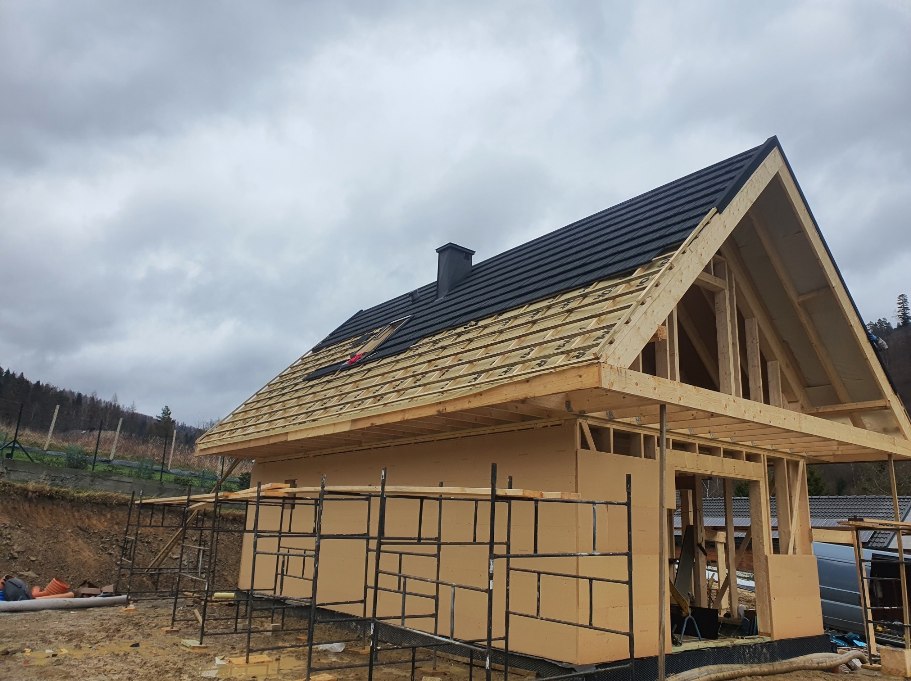 Budowa domu szkieletowego w trakcie: konstrukcja z drewna, częściowo pokryty dach z czarną dachówką, szare niebo w tle. Widoczne rusztowania i teren budowy.