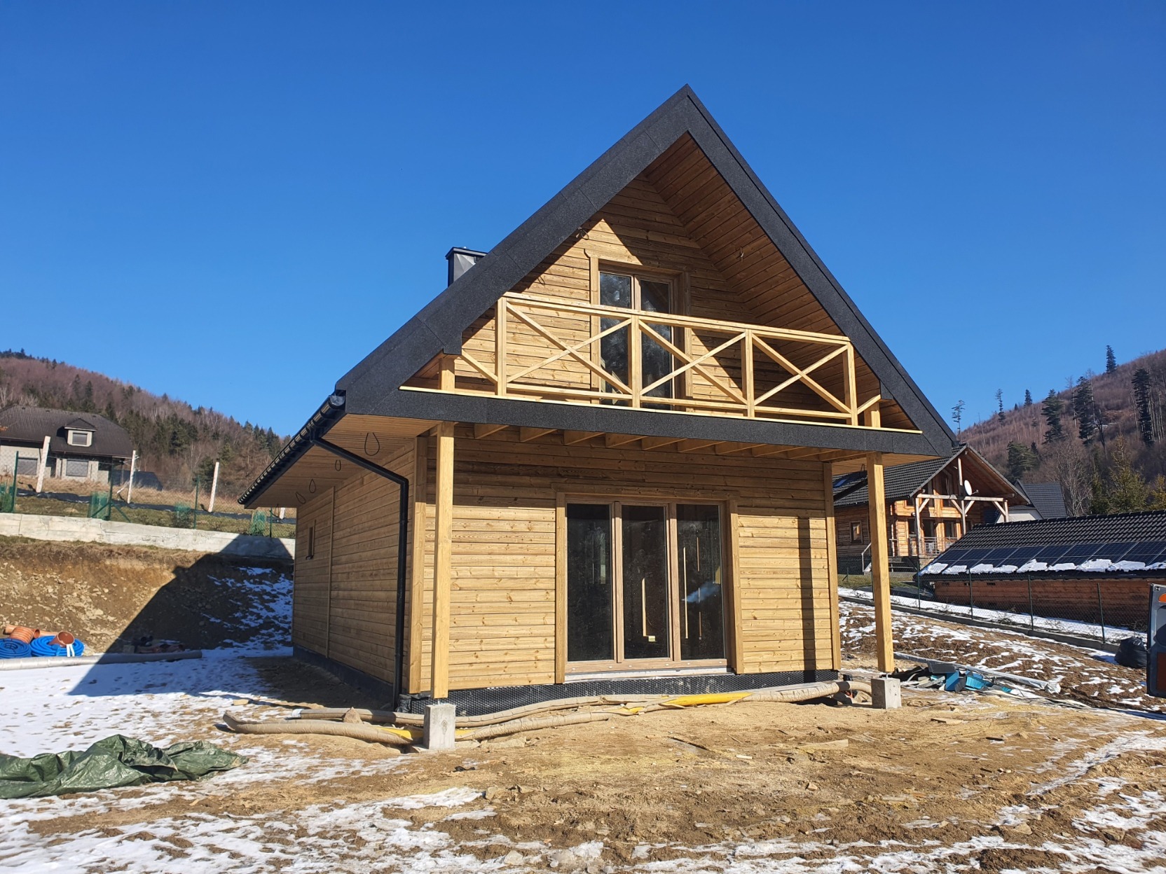 Dwukondygnacyjny dom z drewna w stylu góralskim z balkonem, na tle gór i błękitnego nieba, częściowo ośnieżony teren budowy.