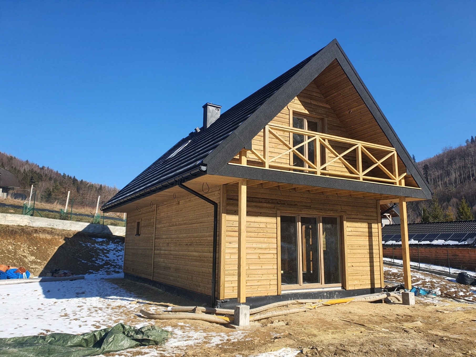 Jasny, dwukondygnacyjny dom szkieletowy z balkonem i czarnym dachem na tle błękitnego nieba i zalesionych wzgórz. Widoczne elementy wykończeniowe i teren wokół budynku.