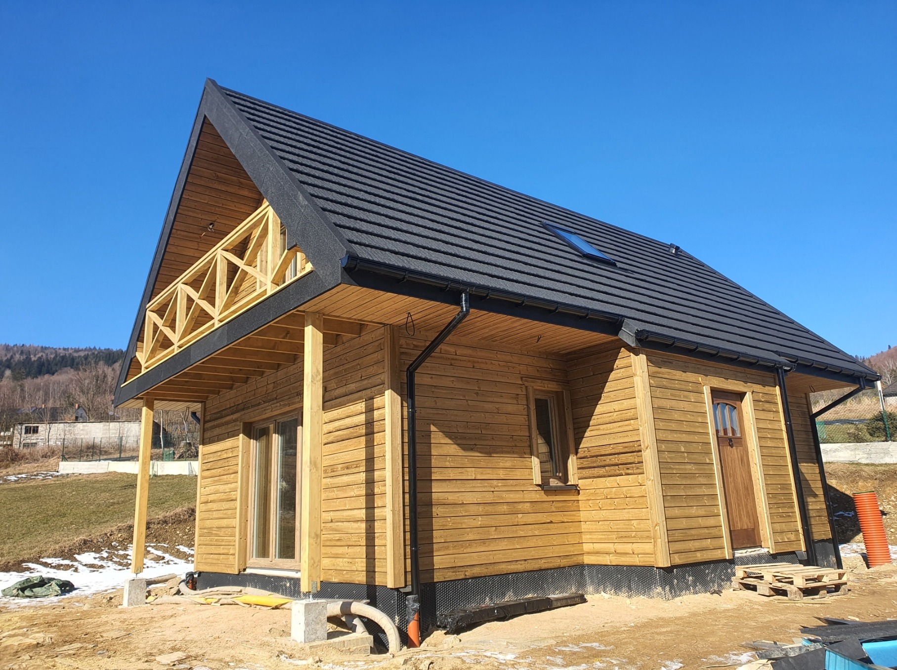 Drewniany dom z czarnym dachem i balkonem z drewnianą balustradą, widok z dołu na tle błękitnego nieba. Wokół domu widać resztki śniegu i materiały budowlane.