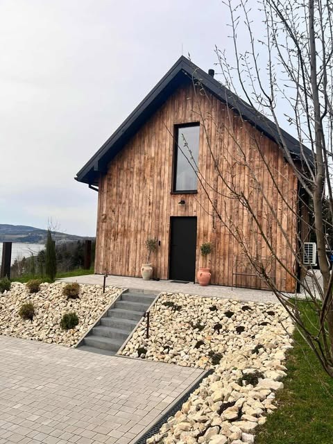 Nowoczesny dom z drewnianą elewacją, czarnym dachem i oknami. Schody prowadzą do czarnych drzwi wejściowych. Ogród z kamieniami i roślinami. Widok na jezioro w tle.